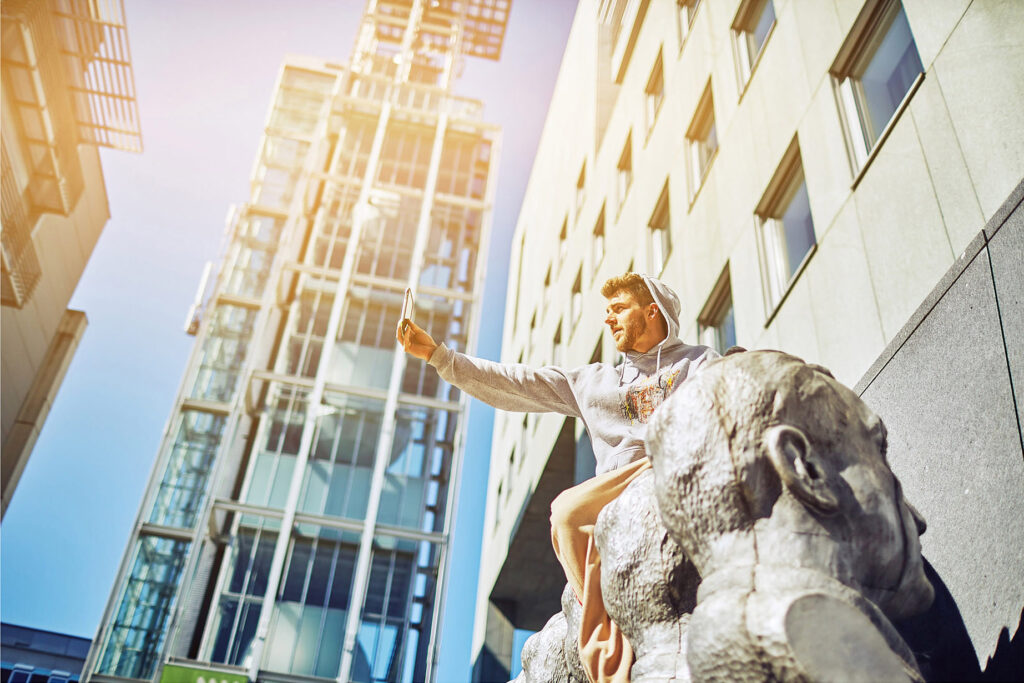 Junger Mann in Hoodie macht ein Selfie auf einer Skulptur vor moderner Glasfassade – urbane Lifestyle-Szene fotografiert von Atelier Schulte.