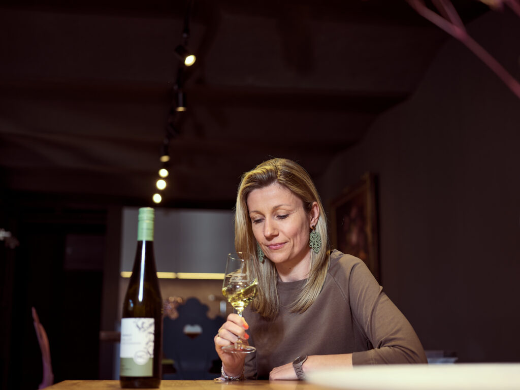Businessportrait einer Winzerin sitzend am Holztisch im Verkostungsraum, aufgenommen von Atelier Schulte.