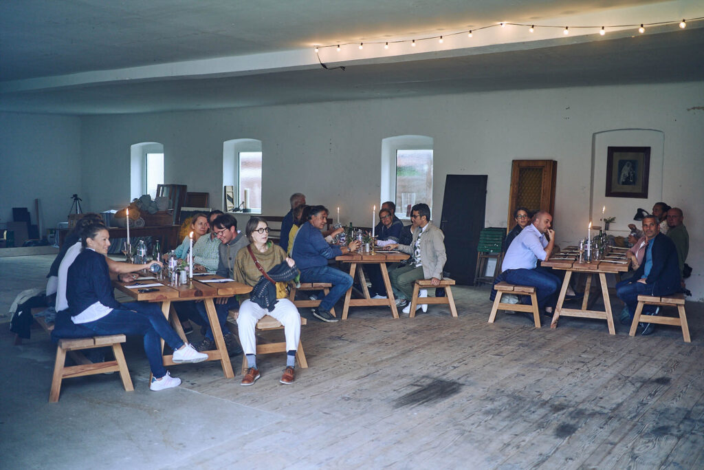 Gäste sitzen an Holztischen beim Abendessen während Essen vor Ort am Wagram in Niederösterreich. Kulinarische Reportagefotografie mit Atmosphäre und Lichtstimmung, Atelier Schulte.