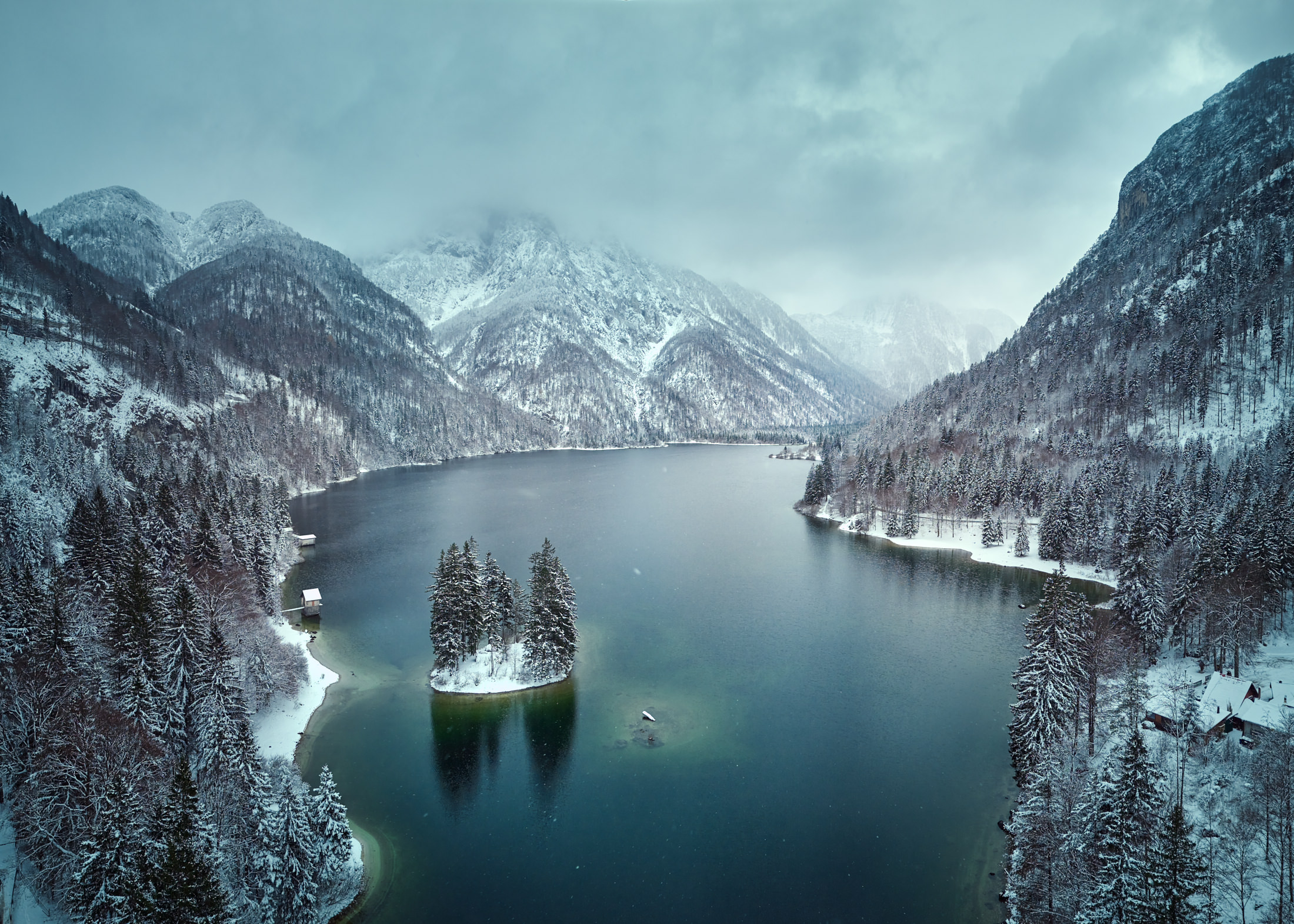 Drohnenaufnahme des winterlichen Lago di Predil mit verschneiter Insel, dunklem Wasser und dramatischer alpiner Bergkulisse – professionelle Landschaftsfotografie von Atelier Schulte