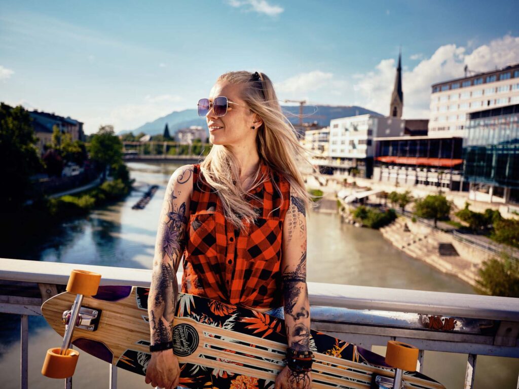 Frau mit Skateboard steht auf einer Brücke in Villach – urbaner Lifestyle-Moment mit Fluss, Sommerlicht und entspannter Atmosphäre in Kärnten