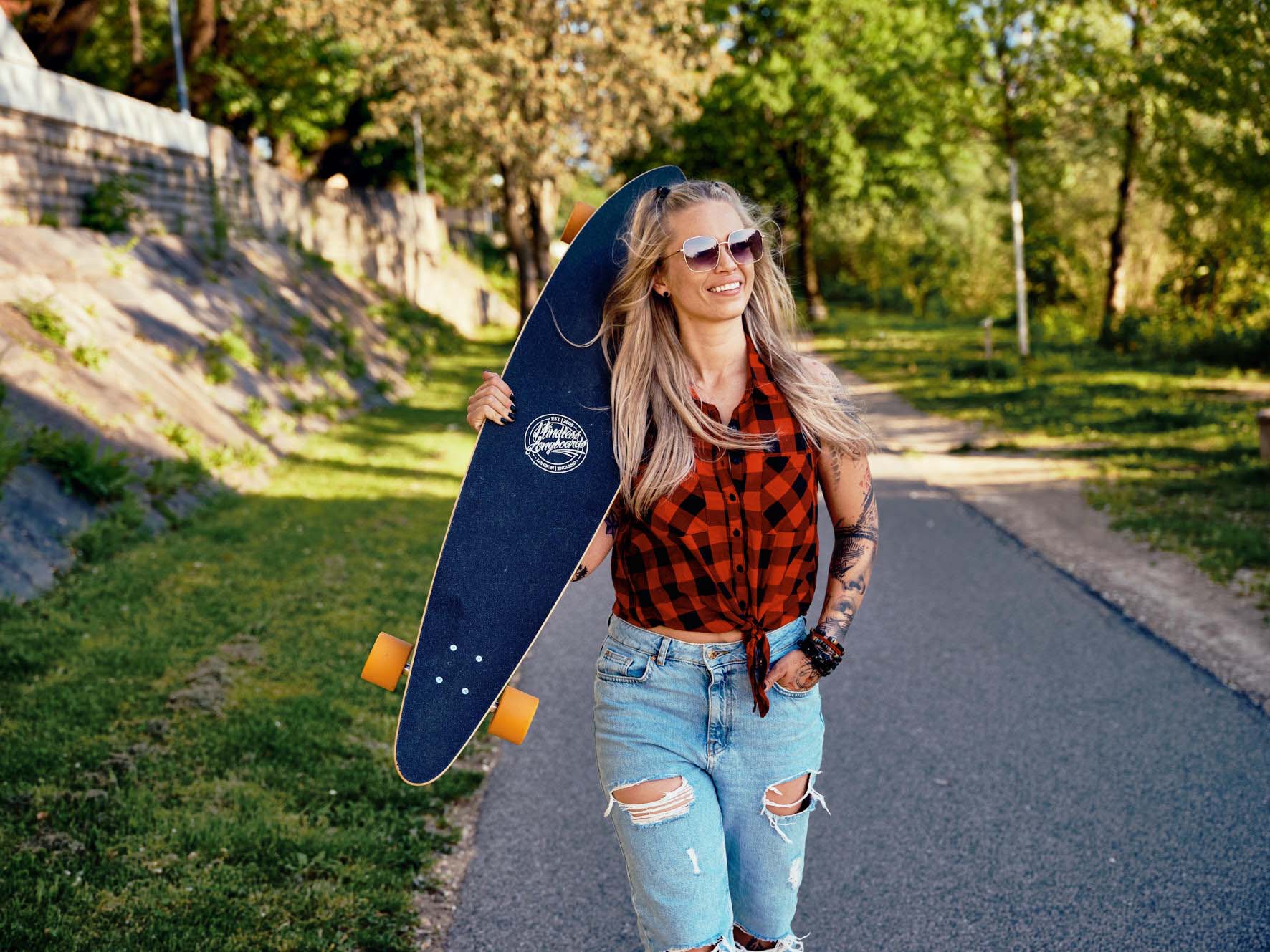 Frau mit Skateboard geht entspannt auf grünem Weg in Villach – sommerlicher Lifestyle-Outdoor-Moment mit Natur, Bewegung und Leichtigkeit in Kärnten