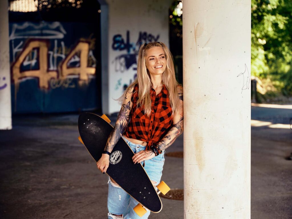 Lifestyle-Outdoor-Fotografie in Villach, Kärnten: urbanes Portrait einer jungen Frau mit Skateboard, entspannt an einer Betonsäule, natürliches Licht und ungezwungene Atmosphäre