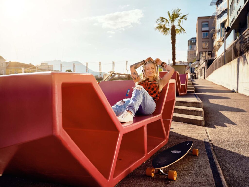 Lifestyle-Outdoor-Fotografie aus Villach in Kärnten: junge Frau entspannt auf urbaner Sitzskulptur, Skateboard daneben, Sommerlicht und lockere Atmosphäre