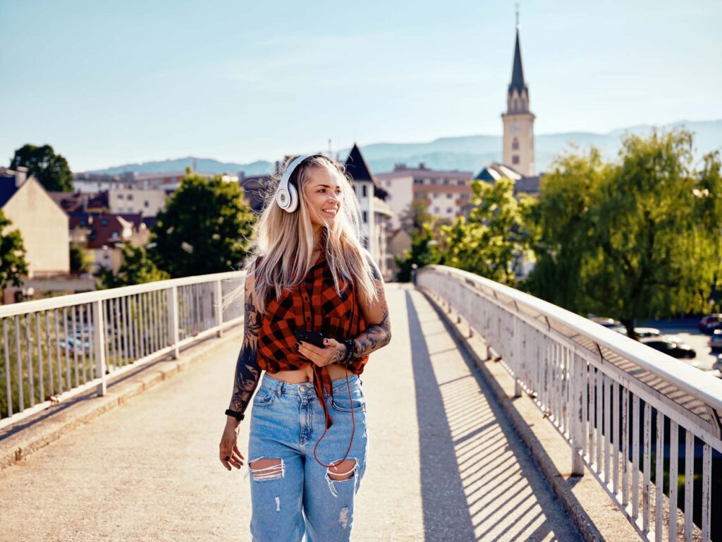 Frau mit Kopfhörern geht über eine Brücke in Villach – urbane Bewegung mit Sommerlicht, entspanntem Lifestyle und Stadtkulisse in Kärnten