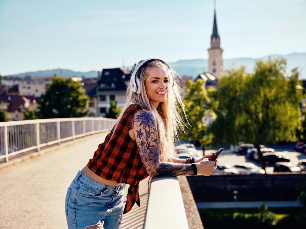 Frau mit Kopfhörern lehnt auf einer Brücke in Villach – urbaner Lifestyle-Moment mit Sommerlicht, Ruhe und Blick über die Stadt in Kärnten