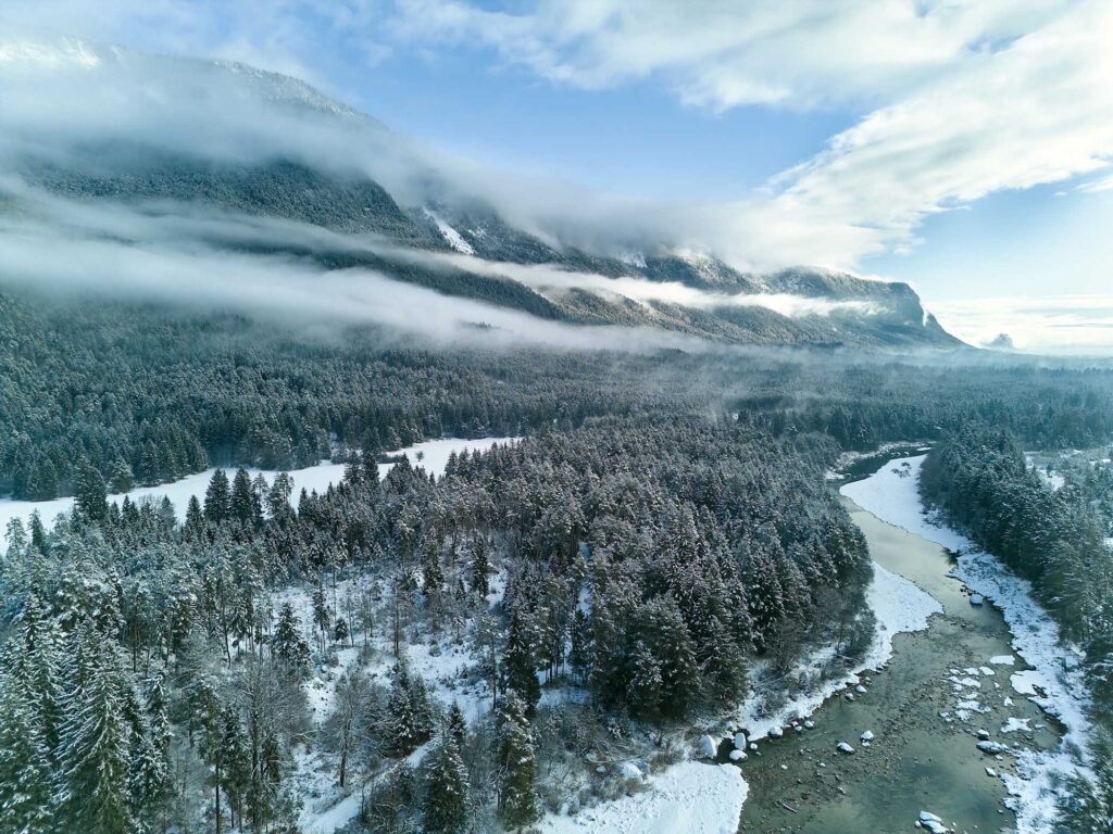Winterlicher Blick über das Tal der Schütt mit verschneiten Wäldern, Flusslauf und tief liegenden Nebelbändern vor der Bergkulisse – fotografiert von Atelier Schulte