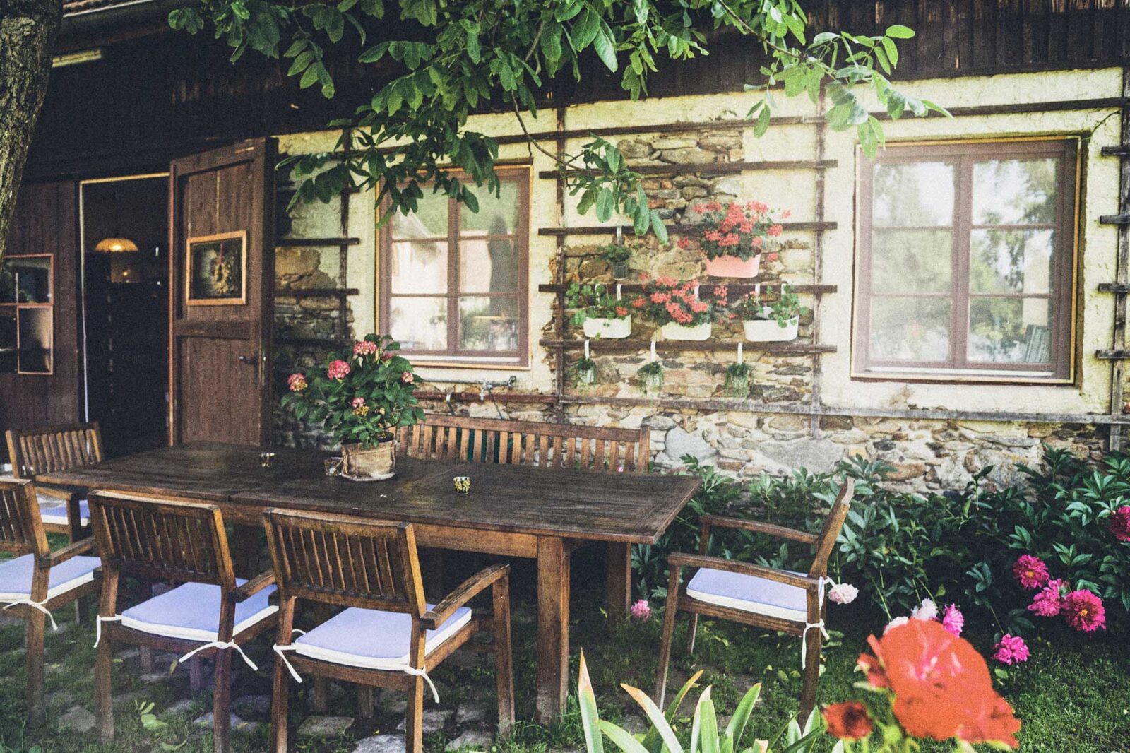 Sommerliche Hofterrasse in der Wachau mit Holztisch, Steinmauer und blühenden Pflanzen – analoge Architekturreportage von Atelier Schulte mit Fokus auf Licht, Atmosphäre und Außenräume.