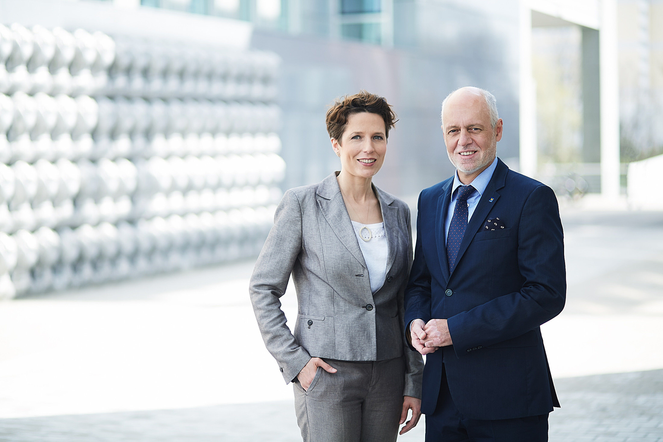 Zwei Personen in Businesskleidung vor einem modernen Gebäudekomplex – professionelles Businessportrait im Außenbereich.
