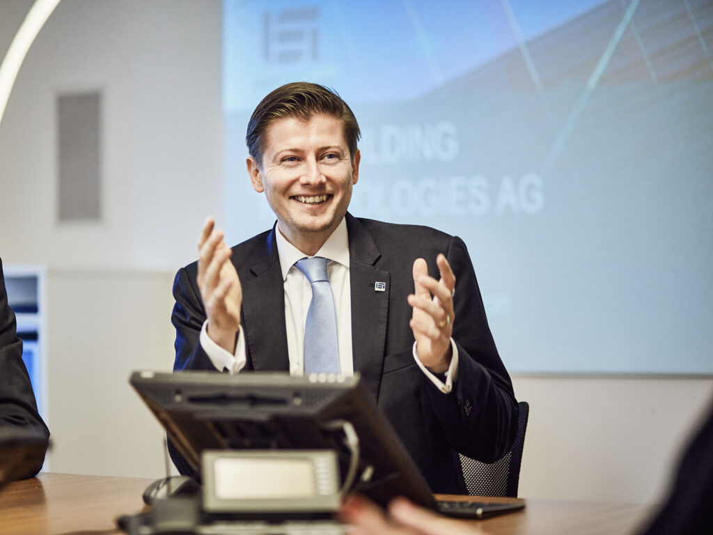 Geschäftsführer spricht gestikulierend in einer Besprechungssituation vor Präsentationsfläche, aufgenommen in professionellem Business-Umfeld.