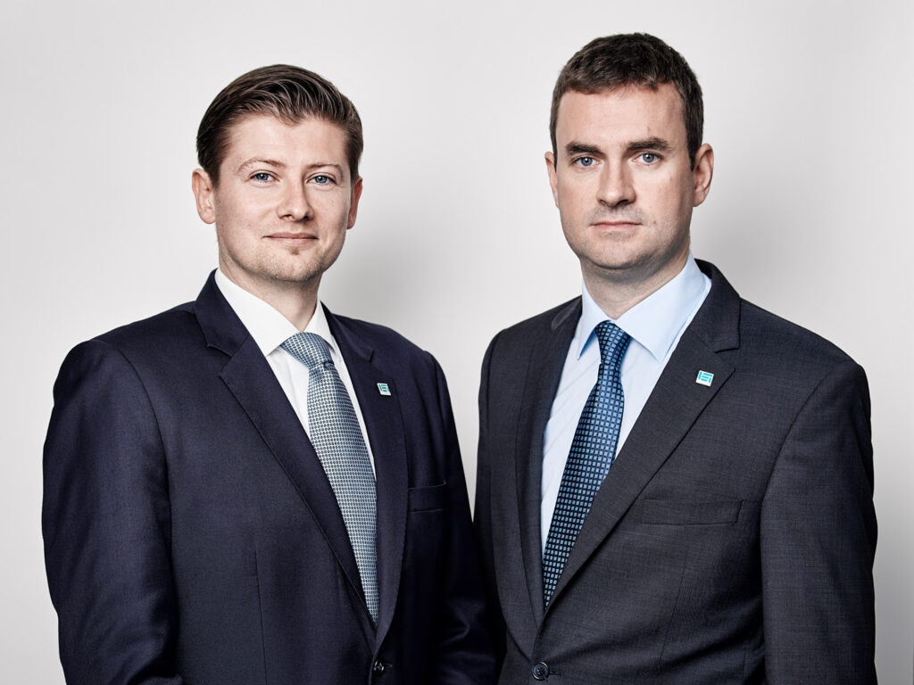 Zwei Führungskräfte stehen nebeneinander vor neutralem Hintergrund, aufgenommen als professionelles Businessportrait.