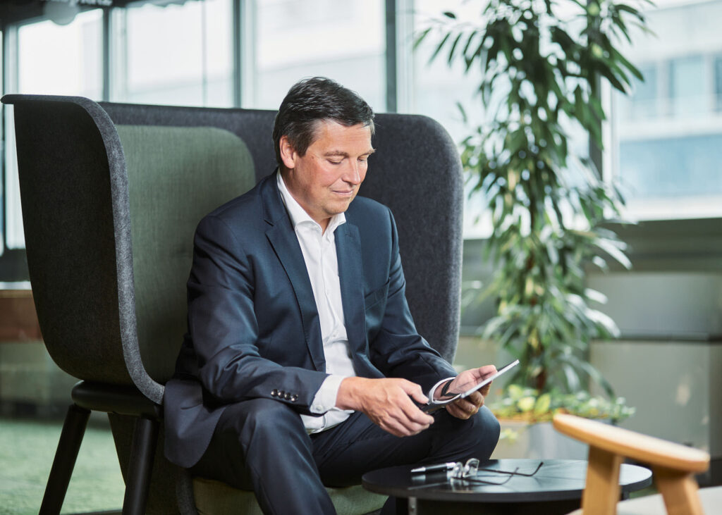 Businessportrait eines Managers im modernen Bürobereich – sitzend im Lounge-Sessel, mit Tablet und Brille in der Hand. Beispiel für authentische Mitarbeiterfotos und typische Kostenfaktoren in der Unternehmenskommunikation.