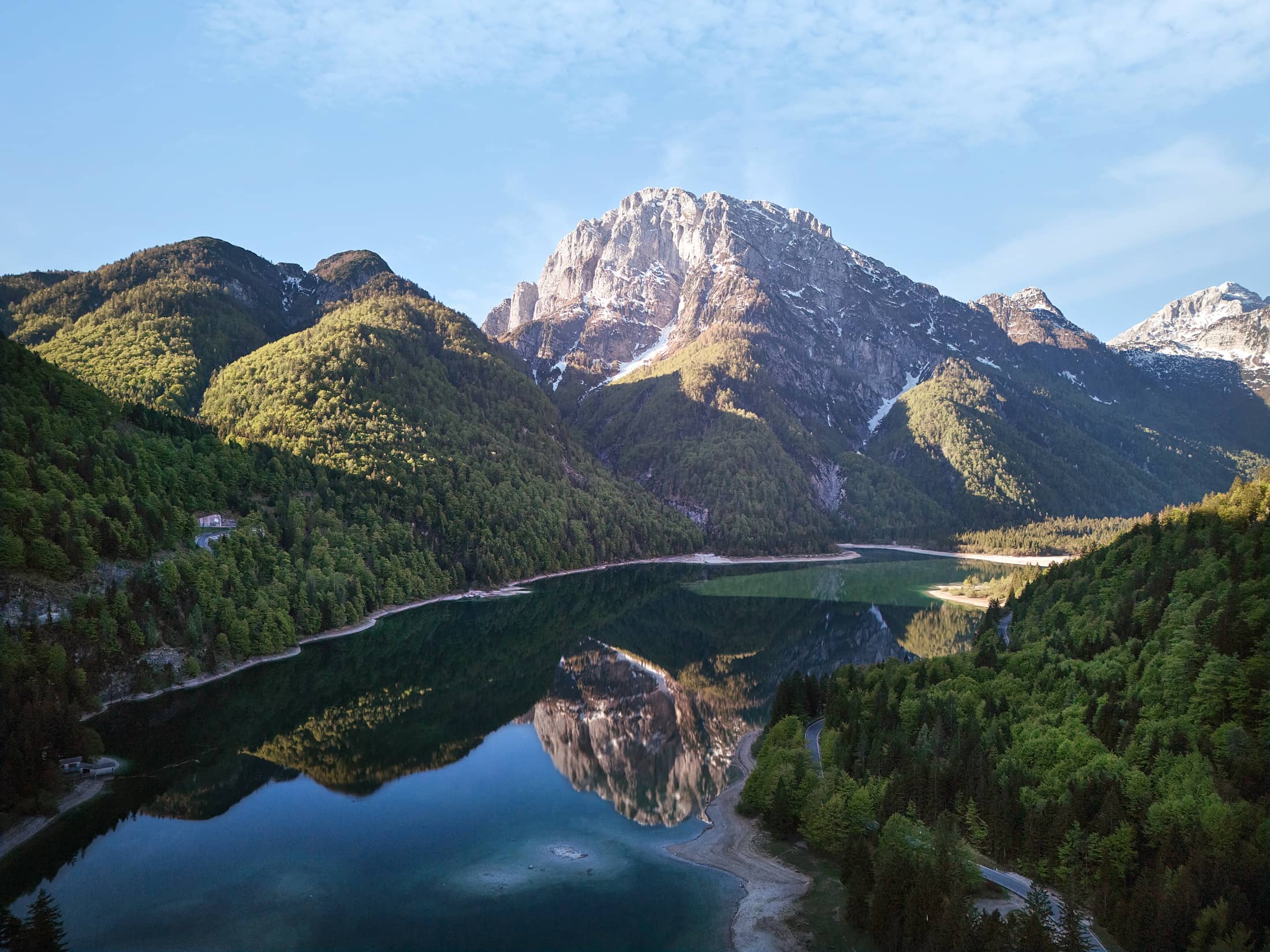 Drohnenaufnahme eines Bergsees im Alpen-Adria-Raum mit ruhiger Spiegelung, Frühlingslicht und kurviger Straße – professionelle Landschaftsfotografie von Atelier Schulte in Österreich.